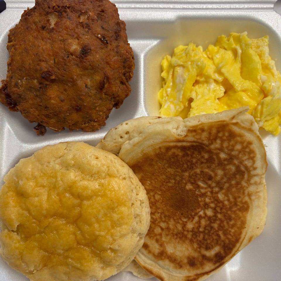 A breakfast platter with a fried patty, scrambled eggs, a biscuit, and pancakes in a styrofoam container.